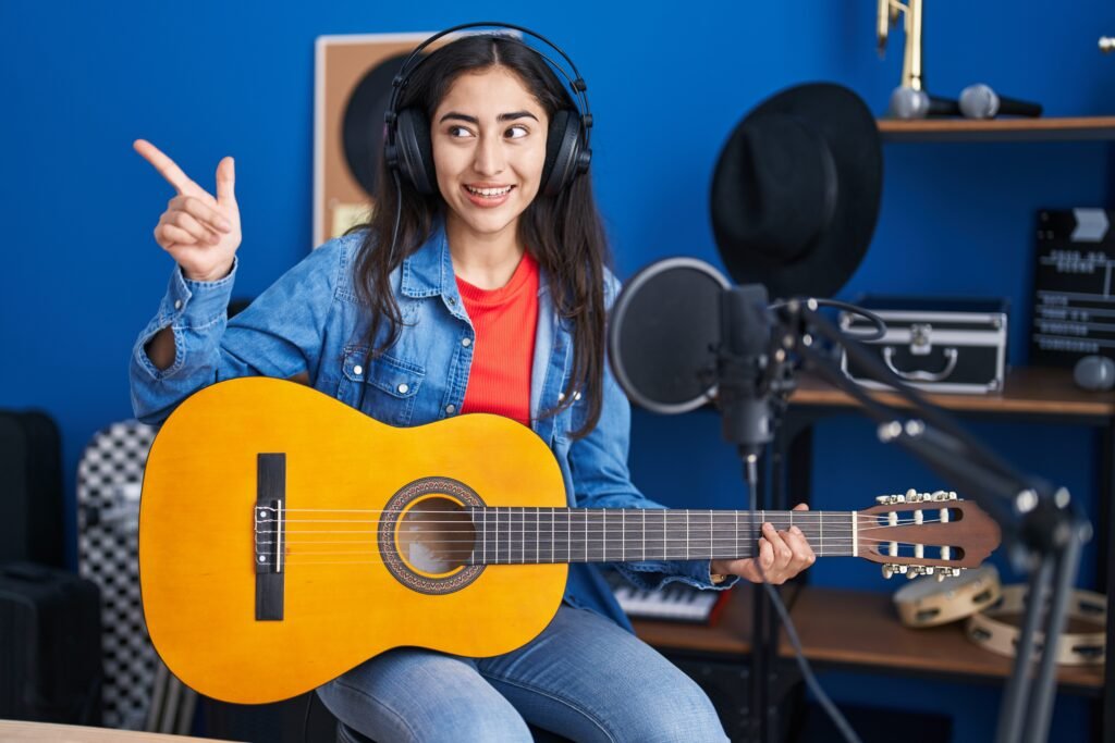 young teenager girl playing classic guitar at music studio pointing thumb up to the side smiling happy with open mouth